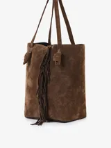 Bolso tipo bucket confeccionado en gamuza color marrón, con flecos aplicados a mano en el centro que aportan movimiento. Posee manijas para llevar al hombro, un sobre interno y una correa larga de terciopelo con detalles en gamuza.
