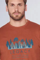 Remera de manga corta con cuello redondo, color naranja terracota. Presenta un estampado frontal que incluye un bosque de pinos en tonos azules y el texto 'LEGACY' junto a una frase inspiradora.