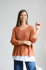 Sweter de punto color terracota con diseño amplio y escote en V con detalle de vivo plateado. Presenta mangas largas y un diseño de doble capa con una base blanca visible en el ruedo.