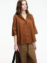 Camisa de corte holgado y diseño minimalista, confeccionada en algodón color marrón. Presenta cuello camisero, cierre frontal con botones y mangas tres cuartos.