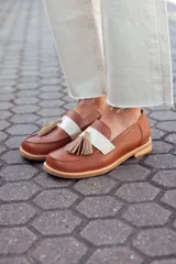 Mocasines de cuero color tostado, con detalle de antifaz en cuero crudo y borlas decorativas. Hechos a mano en Uruguay.