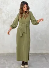 Vestido largo color verde militar, con cuello redondo, mangas largas abullonadas con puños elásticos y detalle de lazo anudado en la cintura. Presenta una abertura lateral en la falda.