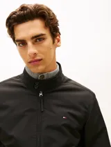 Campera tipo Harrington de Tommy Hilfiger en color negro, de corte amplio y tejido repelente al agua. Cuenta con cuello levantado, cierre frontal de cremallera con botón, bolsillos de ojal en la cintura, y puños y bajo de punto canalé. Presenta el logo de la marca bordado en el pecho.