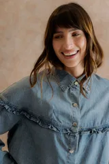 Camisa de denim liviano con calce relajado, cuello camisero clásico y cierre frontal con broches metálicos. Presenta un canesú con volado en el delantero y la espalda, mangas largas con puños abotonados y terminación inferior redondeada.