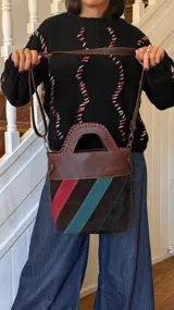 Matera de cuero con diseño estructurado, asa superior rígida y correa larga ajustable. El cuerpo presenta un diseño de franjas diagonales en gamuza de colores marrón, rojo, turquesa y negro.