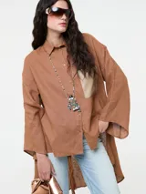 Camisa larga de lino color marrón, con corte oversize, mangas anchas y bolsillo frontal de cuero sintético en tono contrastante. Presenta cierre frontal con botones y diseño de bajo irregular.