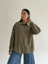 Camisa oversize de color marrón con cuello camisero y puños con detalle de bordado festoneado en color blanco. Presenta cierre frontal de botones y mangas largas.