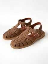 Sandalias estilo cangrejera color marrón, confeccionadas en cuero con diseño trenzado en la parte delantera, tira ajustable con hebilla metálica en el tobillo y suela plana.