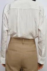 Camisa fluida de corte oversize confeccionada en mezcla de viscosa, con diseño de rayas verticales finas en tono beige sobre fondo blanco. Presenta cuello solapa, cierre frontal con botones, manga larga con puños y un bolsillo de plastrón en el pecho.
