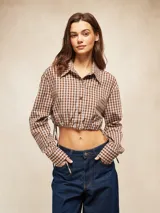Camisa de manga larga, cuello camisero y botones frontales, con estampado a cuadros marrones y blancos. El ruedo inferior es ajustable con cordones laterales, creando un efecto *cropped* y fruncido.