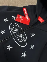 Buzo con cierre frontal, color negro, con estampado gráfico de estrellas y logo en el pecho. Presenta capucha, bolsillos tipo canguro y puños elásticos.
