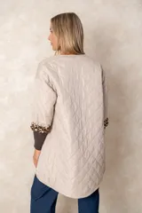 Campera acolchada de corte amplio y largo midi, color beige con diseño capitoneado. Presenta mangas tres cuartos con puños vueltos que revelan un interior estampado con motivo animal print. Diseño sin cuello y cierre frontal oculto.