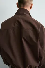 Cazadora técnica de corte boxy fit en color marrón, con cuello subido, cierre frontal de cremallera, bolsillos laterales y uno frontal con cierre, y acabados elásticos en puños y ruedo.