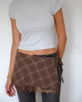 Falda tipo kilt de diseño envolvente, confeccionada en tela a cuadros marrones con detalle de encaje en el ruedo y cierre lateral con lazos.