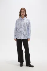 Camisa de algodón con diseño de rayas verticales finas en color celeste y blanco, con apliques florales bordados en relieve. Presenta cuello camisero, cierre frontal con botones y mangas largas con puños abotonados.