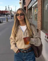 Sweater de punto color beige con diseño de trenzas, cierre frontal completo, cuello alto y mangas largas con puños ajustados. Presenta un corte ligeramente corto.
