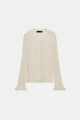 Chaqueta de punto canalé en color beige con hilo metalizado, cuello redondo, manga larga y cierre frontal con botones. Presenta detalles de volantes en la tapeta de los botones, el ruedo y los puños.