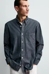 Camisa de corte relajado confeccionada en tejido de algodón denim, con cuello solapa, manga larga con puños abotonados y bolsillo plastrón en el pecho. Presenta cierre frontal de botones y bajo con aberturas laterales.