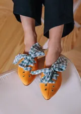 Bailarina de piel natural color naranja con acabado liso, punta redondeada y suela plana. Presenta detalles de ojales metálicos plateados a lo largo del lateral y un lazo desmontable de tela a cuadros vichy en tonos verde agua y blanco sobre el empeine.