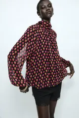 Camisa de cuello subido y manga larga con puños elásticos. Presenta un diseño de lunares en tonos fucsia y amarillo sobre fondo negro, con detalle de pliegues en el frente y cierre de cremallera oculta en la espalda.