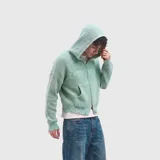 Buzo tejido de punto color verde menta con capucha, cierre frontal completo y bolsillos tipo canguro. Presenta un diseño con efecto desgastado en los puños y el ruedo, y detalles de textura en el pecho.
