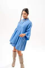 Vestido corto azul, estilo camisero, con cuello camisero, cierre frontal con botones y mangas acampanadas con volados. Presenta un diseño holgado con cortes fruncidos en el cuerpo.