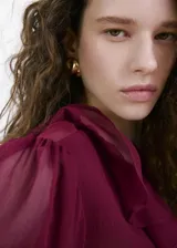 Blusa bordó de tejido fluido y semi transparente, con escote en V pronunciado, mangas largas abullonadas con volantes en los puños y detalles de volantes en el cuello y el ruedo.