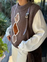 Chaleco tejido de punto en color chocolate, con cuello redondo y diseño sin mangas. Presenta un aplique frontal decorativo compuesto por tres figuras en forma de rombo en tonos tierra, bordeadas con hilo crudo.