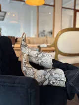 Botas de caña media con estampado de piel de serpiente en tonos beige y marrón. Diseño estilo cowboy con punta cuadrada, detalle de hebilla y apliques metálicos.