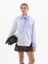 Camisa de poplín con diseño de rayas verticales en tonos azul y blanco. Presenta un cuello prolongado que se anuda en forma de lazo frontal, mangas largas con puños anchos y corte holgado.