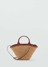Bolso tipo cesta de fibras naturales con diseño en forma de abanico. Presenta detalles en cuero sintético marrón en el borde superior, asas cortas de mano y una correa bandolera larga ajustable. Incluye cierre de botón magnético y forro interior.