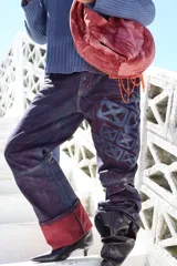 Pantalón de denim de algodón con silueta amplia y construcción estructurada. Presenta un acabado artesanal con efecto de lavado irregular en tonos azul y violeta, destacando un estampado geométrico en una de las piernas y un ruedo con vuelta en color rojo contrastante.