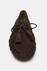 Mocasín de serraje color marrón, con detalle de fruncido en la puntera y lazo con borlas decorativas en el empeine. Presenta una suela plana de 1,5 cm.