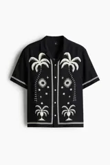 Camisa de manga corta de H&M, con corte relajado, cuello abierto estilo resort, tapeta frontal con botones y bajo recto. Diseño estampado en color blanco sobre base negra con motivos de palmeras, soles y estrellas.