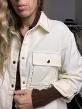 Sobre camisa color manteca con costuras decorativas en zigzag de color oscuro. Presenta cuello clásico, cierre frontal con botones metálicos a presión, mangas largas con puños abotonados y un bolsillo frontal con solapa.