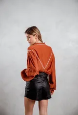 Camisa de tela texturizada color terracota, con mangas largas abullonadas y detalle de bordado blanco en el canesú delantero. Presenta cierre frontal con botones.