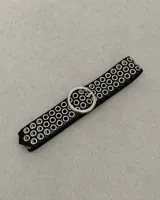 Cinto ancho de cuero negro decorado con múltiples filas de ojalillos metálicos plateados y hebilla circular grande.