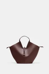 Bolso tipo shopper con forma de capazo, color marrón oscuro, confeccionado en material liso con pespuntes visibles en el cuerpo. Posee doble asa de mano y un asa bandolera ajustable. Incluye un bolso interior extraíble que se cierra mediante fruncido y un cierre principal con botón imantado.