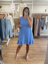 Vestido corto sin mangas, color gris topo, con escote redondo y detalle de volado en el ruedo. El tejido presenta una textura arrugada característica del lino.