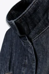 Cazadora de denim color azul oscuro, con cuello solapa, mangas largas tipo murciélago y bolsillos de vivo en el delantero. Presenta un bajo elástico ajustable con cordones y cierre frontal con cremallera y botones a presión ocultos.