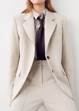 Blazer color beige con solapas de muesca, cierre frontal de dos botones oscuros, bolsillos con solapa en los laterales y un bolsillo ojal en el pecho.