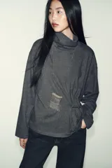 Top de cuello alto y manga larga, confeccionado en tejido de algodón gris melange. Presenta un diseño con frunces laterales ajustados mediante un aplique metálico decorativo.