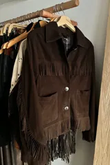 Campera de gamuza color chocolate con diseño de flecos en el pecho, espalda y mangas. Presenta cuello clásico, cierre frontal con botones y bolsillos laterales.