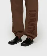 Pantalón deportivo oversize de felpa, color marrón, con cintura elástica y cordón ajustable. Presenta puños acanalados y un parche bordado con el texto "10 DAYS" en el lateral de la pierna.