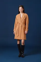 Saco largo tipo trench de color tostado, con diseño de doble botonadura, solapas clásicas y cinturón ajustable en la cintura. Presenta mangas largas con trabillas en los puños y una silueta estructurada y fluida.