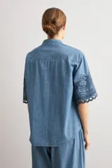 Camisa de denim celeste con cuello mao, botonadura frontal oculta y mangas cortas holgadas que presentan un delicado bordado floral oscuro y dobladillo calado.