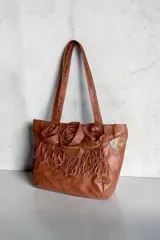 Cartera shopper de cuero color marrón terra, con doble asa de hombro y diseño decorativo de tres flores de cuero y flecos en el frente.