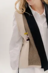 Chaleco abierto de tejido de punto, con diseño de bloques de color verticales en gris claro, negro y gris oscuro. Presenta un cuello ancho tipo chal y un detalle de tres pequeñas aplicaciones bordadas horizontales en tonos verde oliva, naranja quemado y celeste pálido en el frente.