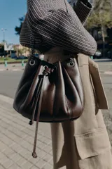 Bolso tipo saca de cuero sintético color marrón oscuro, con diseño fruncido y cierre mediante cordón ajustable. Cuenta con asas de hombro combinadas con cadena metálica y bolsillo interior con cremallera.