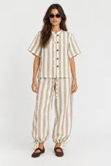 Pantalón a rayas beige y blancas con cintura elástica, bolsillos laterales y traseros, corte holgado con piernas globo y cordón de ajuste en los tobillos.